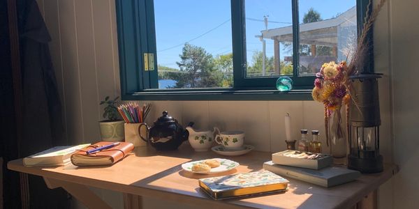 Cozy wooden desk by a sunny open window with tea set and art supplies.