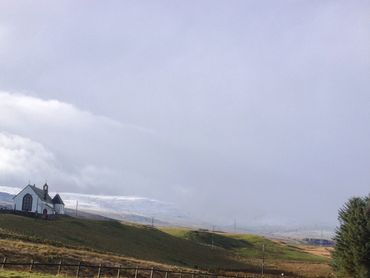 A white church on a green hill with snowy mountains in the background.