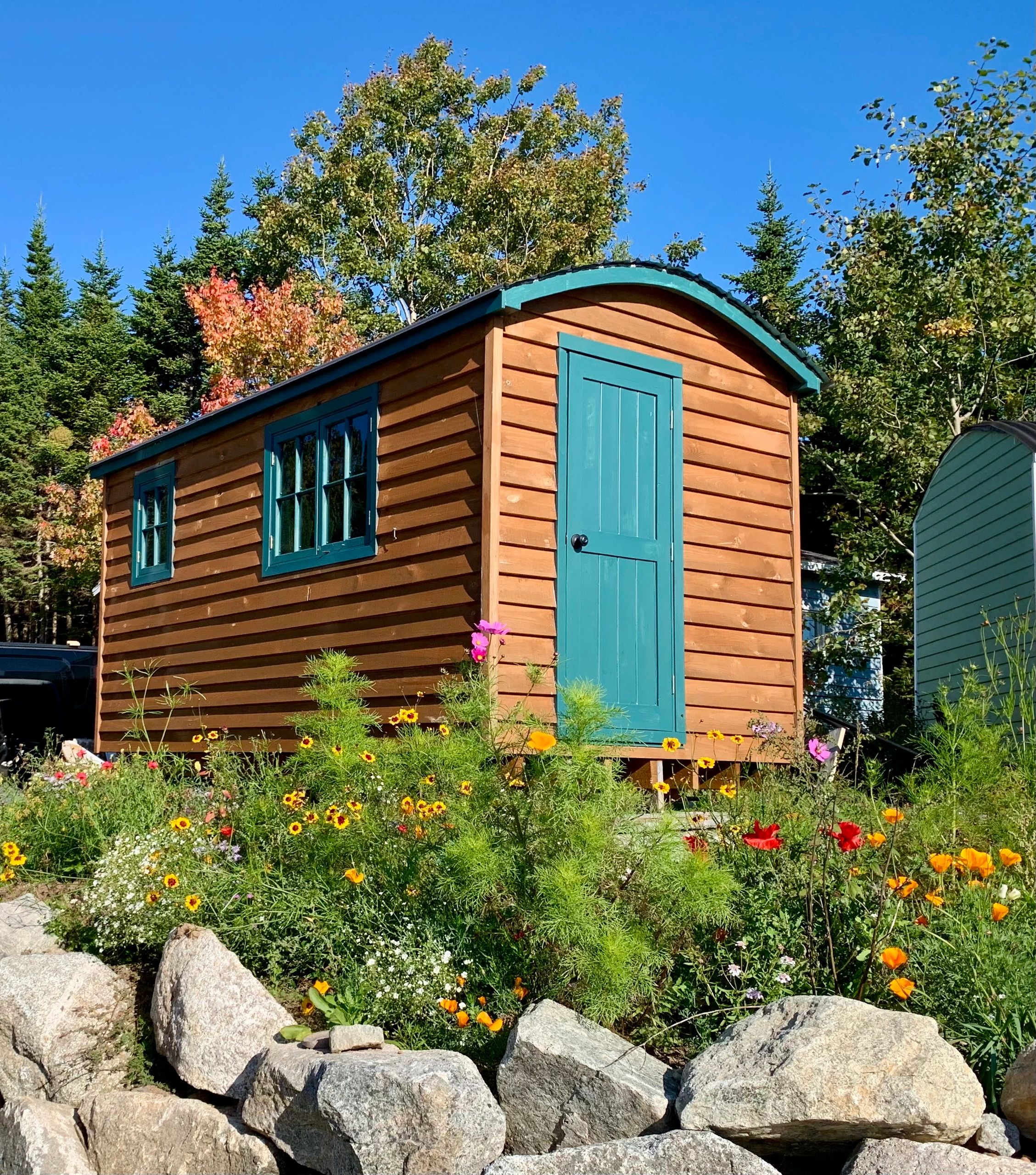 A small wooden cabin with teal door and windows surrounded by colorful flowers and rocks.
