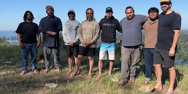 A group of eight men standing outdoors on a grassy hill under a clear sky.