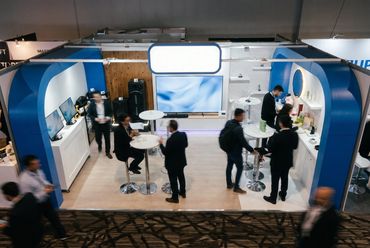 Stand con arcos azules y pantalla, con visitantes reunidos en mesas altas