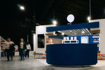 Stand con mostrador azul en una feria, con visitantes recorriendo el pabellón