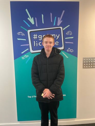 Young man in black jacket stands in front of a #gotmylicense sign.