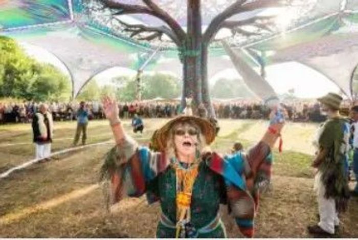 A joyful woman in colorful attire raises her arms at an outdoor festival under a large canopy.