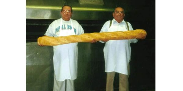 Two bakers holding a giant baguette in a bakery.