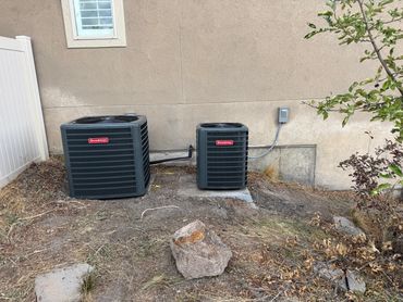 Two Goodman air conditioning units outside a house on a dry patch of ground.