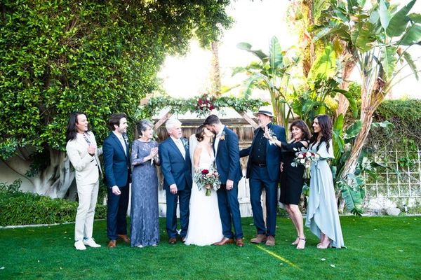 A family wedding photo with the bride and groom kissing. Standing in front of green palm trees.