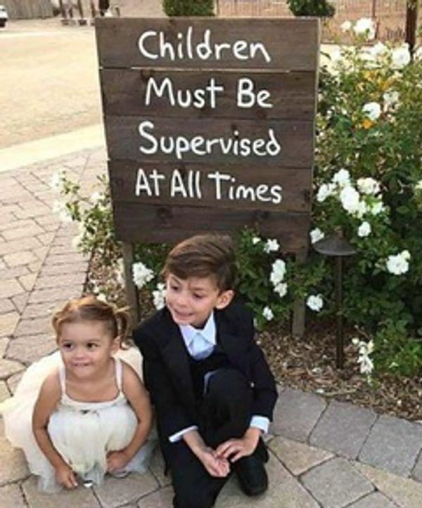 A little flower girl and ring bearer crouched in front of a wooden sign at a wedding.