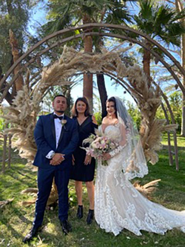 A bride and groom with their minister standing in front of a natural arch and tall palm trees.