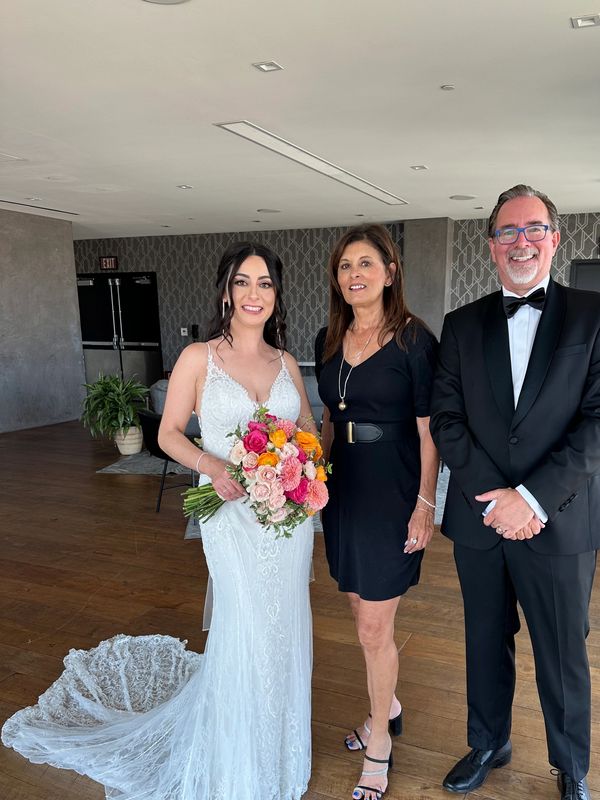 Bride with dark hair in long white gown standing with a man and woman who are both dressed in black.