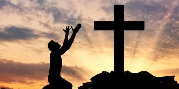 Silhouette of a person praying near a cross at sunset.