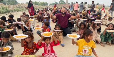 Children and a man holding plates of food, gathered outdoors in a large group.