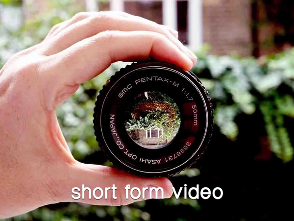 A hand holds a camera lens through which we see a garden scene reflected
