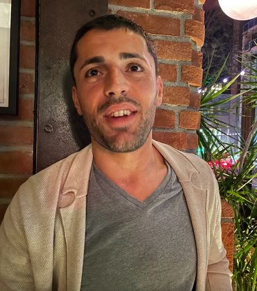 Man with short hair and a light jacket smiling near a brick wall at night.