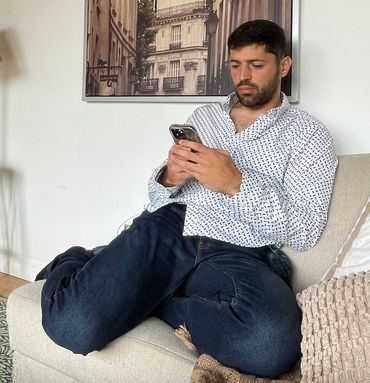 Man sitting on couch using smartphone, wearing a patterned shirt and jeans.