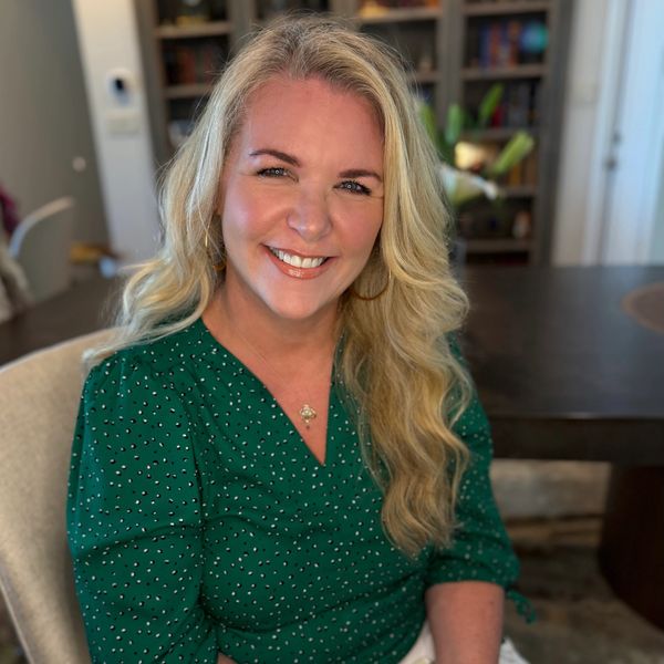 Smiling woman with long blonde hair in a green blouse sitting indoors.