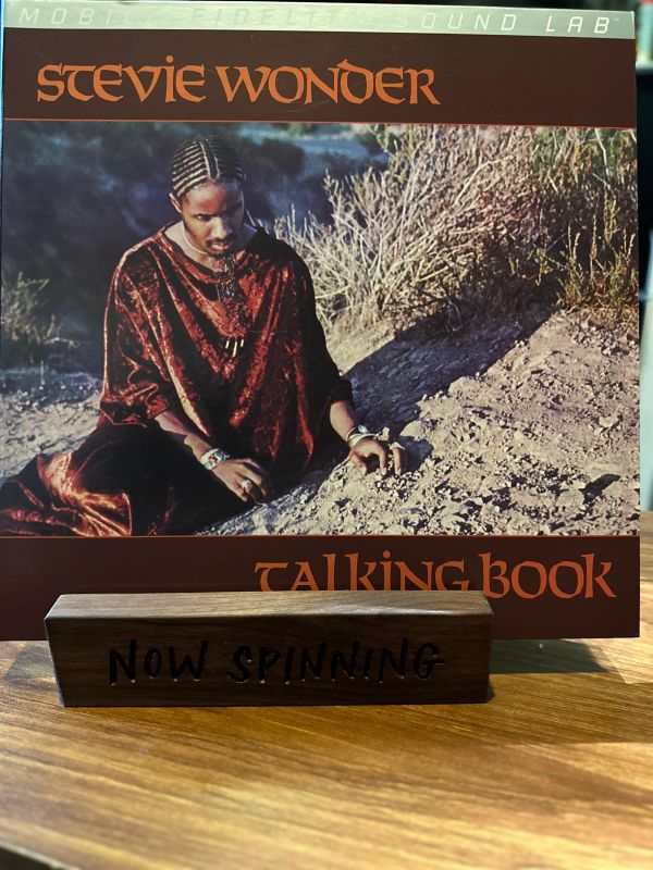 Stevie Wonder's Talking Book album cover with 'Now Spinning' sign on a wooden stand.