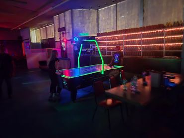 Two people playing air hockey in a dimly lit arcade or game room with neon lights.
