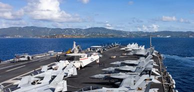 Aircraft carrier deck with fighter jets lined up and ocean in the background.
