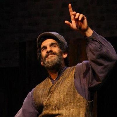 Barry seated in a European peasant costume, bearded, smiling and pointing up at the sky, against a d