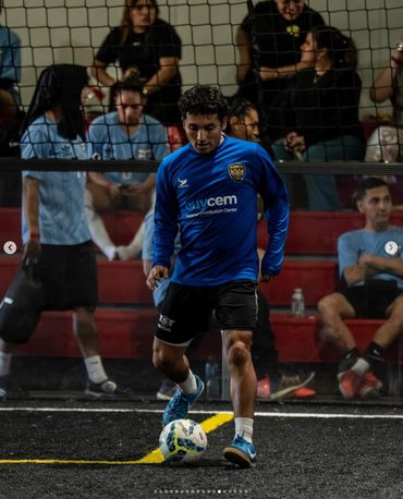 Soccer player in blue jersey controlling the ball on an indoor field with spectators behind the net.