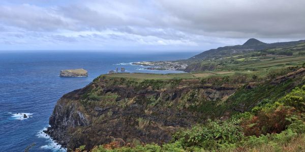 Coastal cliffs with green vegetation overlooking a blue ocean under a cloudy sky.