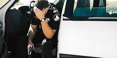A distressed police officer sitting in a patrol car holding his head and a gun.