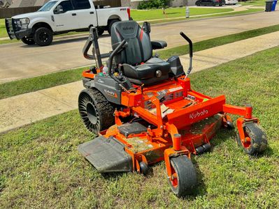 Orange Kubota zero-turn lawn mower parked on a grassy area near a sidewalk.