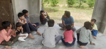 A group of children and a teacher are sitting on the floor in a semi-circle outdoors.