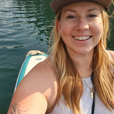 Smiling woman with long hair on a paddleboard in calm water, wearing a cap.