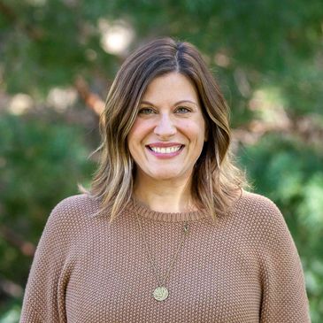 A smiling woman wearing a brown sweater outdoors with greenery in the background.