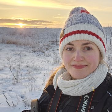 Woman in winter clothes smiling at sunset in snowy landscape.