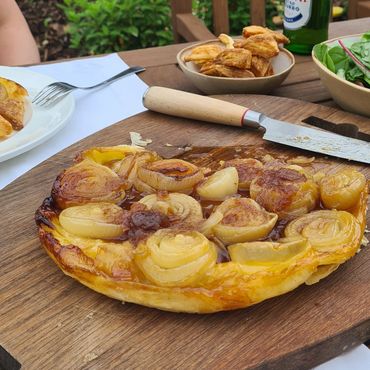 Onion tart on a wooden board with salad, chips, and beer outdoors.
