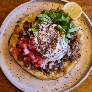 A delicious flatbread topped with seasoned meat, diced tomatoes, onions, greens, and a dollop of yogurt with spices.