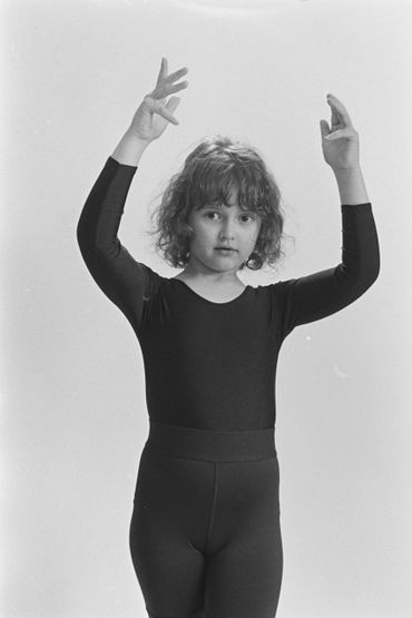 A young girl in a black leotard poses with arms raised gracefully.