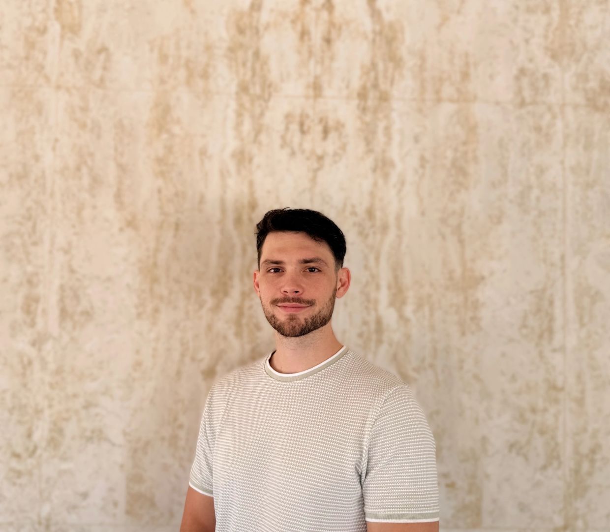 Man with a light-colored shirt standing against a textured wall.