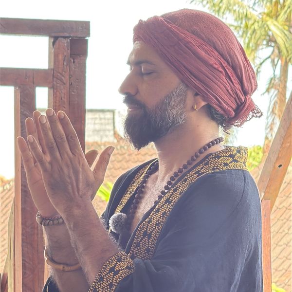 Man meditating with hands together, wearing a red headwrap and a patterned robe.