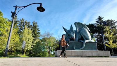 Wide outdoor photo of a person arriving at the U of G, standing beside the large gryphon statue. The