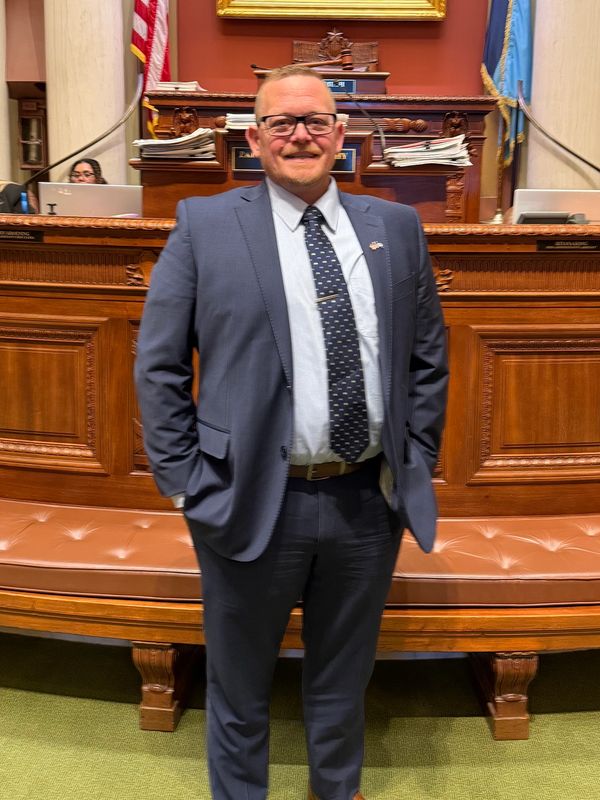 Man in a suit standing in a formal government chamber with a painting behind.