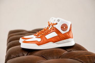 Orange and white CobblerX high-top sneakers displayed on a brown leather bench.