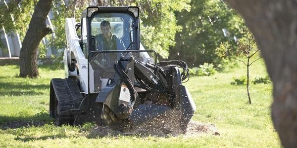 A man operates a compact excavator in a grassy park area.