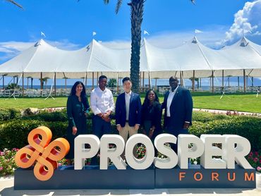 Five professionals posing behind a PROSPER Forum sign outdoors on a sunny day.
