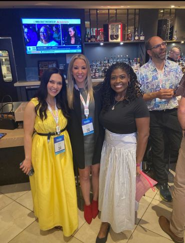 Three women posing and smiling in a bar with a TV showing a WNBA draft event.