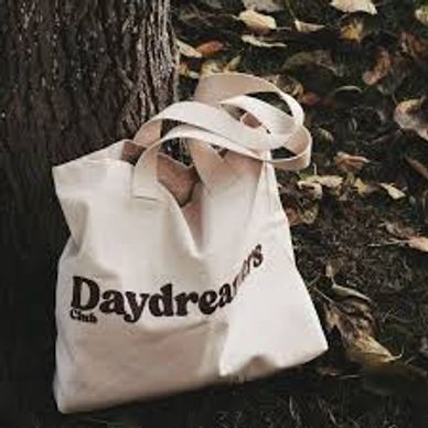 A beige tote bag with "Daydreamers Club" text leaning against a tree.