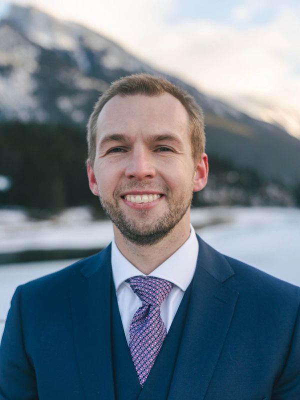 Man in a blue suit smiling outdoors with mountains in the background.