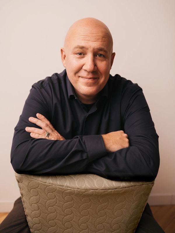 Smiling bald man in black shirt leaning on chair.