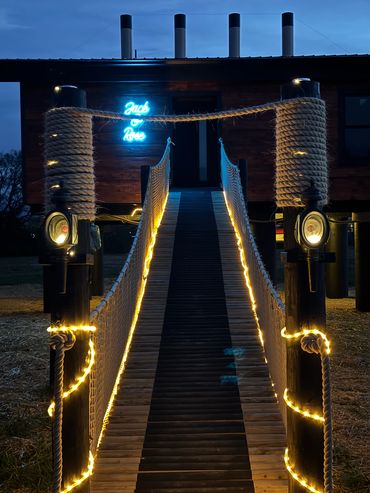 Custom log-rope bridge w/ rope lights, antique carriage lamp lights w/ smokestacks in background.