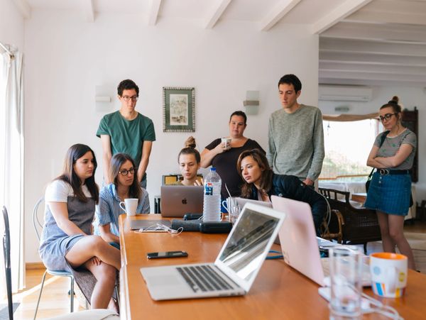 A group of young people collaborating around a table with laptops and drinks.