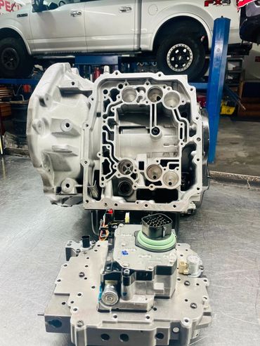 Disassembled vehicle transmission on a workshop table with a truck on a lift in the background.