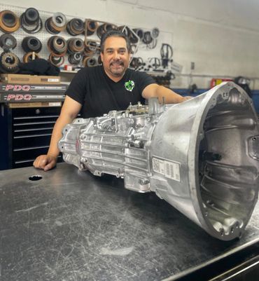 Man smiling behind a car transmission on a metal workbench.